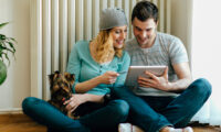 Young Couple Sitting On Floor in a living room at home and shopping online with digital tablet. Smiling woman with hat holding credit card, and man holding tablet and their cute dog on the lap. Their dog is female puppy of Yorkshire Terrier.